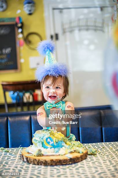 baby boy wearing party hat smashing first birthday cake at table - cake smashing stock pictures, royalty-free photos & images