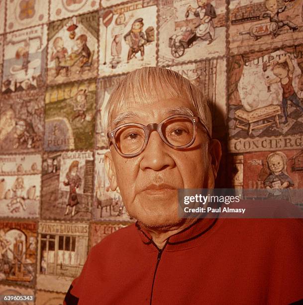 Artist Foujita poses in front of a series of small paintings of places and people. | Location: Artist's home in France.