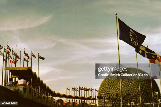 Guests travel on a monorail past the flags of multiple countries, with the Spaceship Earth ride visible in the background, at the Disneyworld park...