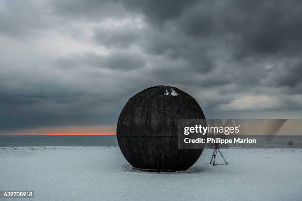 a large black wooden sphere lies on a snow covered shoreline with storm clouds and photographer's tripod - installation art stock pictures, royalty-free photos & images