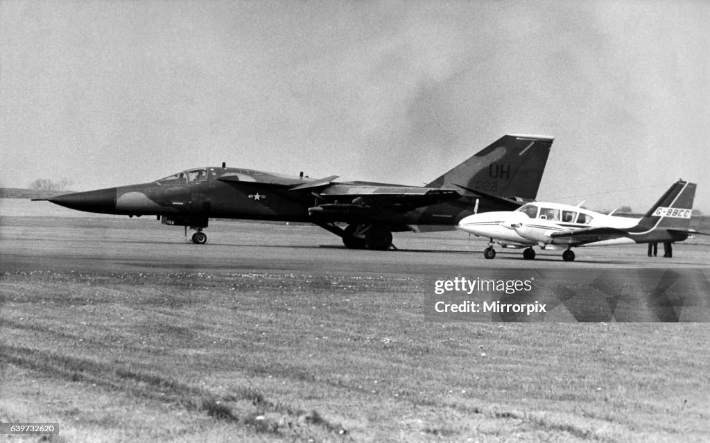 A USAF General Dynamics F-111 Aardvark sits on an airfield next to twin engined civil aircraft