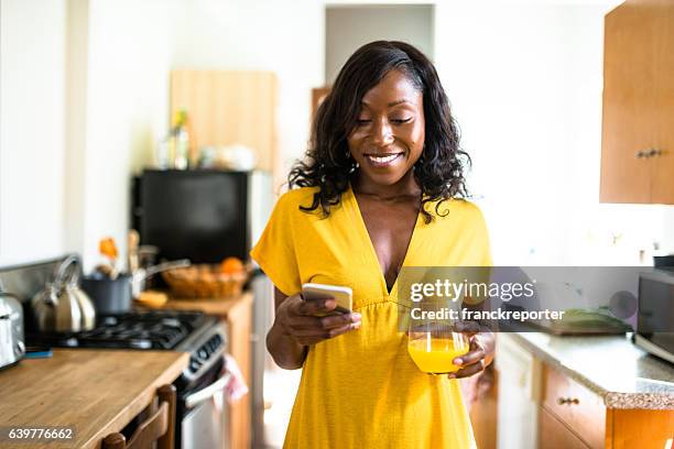 african young woman smiling drinking orange juice - woman drinking phone kitchen stockfoto's en -beelden