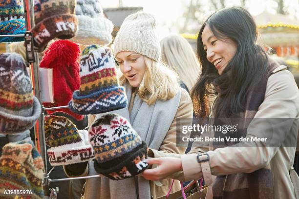female friends are looking at woolen hats at outdoor christmas market stall. - market stall stock pictures, royalty-free photos & images