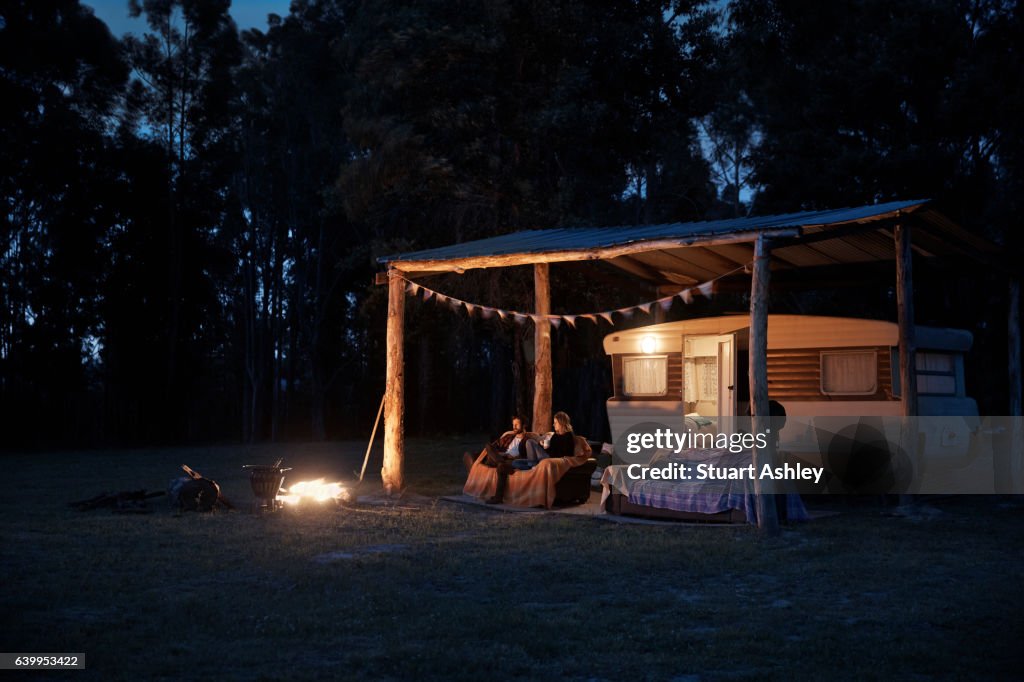Man and woman couple sitting by caravan and fire at night