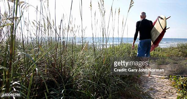 senior male surfer walking on sandy path towards the ocean carrying surfboard. - st augustine florida fotografías e imágenes de stock