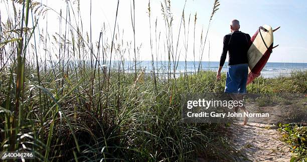 senior male surfer walking on sandy path towards the ocean carrying surfboard. - long grass bildbanksfoton och bilder