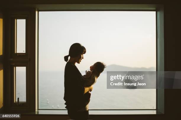 mother hugging baby girl at home in front of window - korean baby girl stock-fotos und bilder