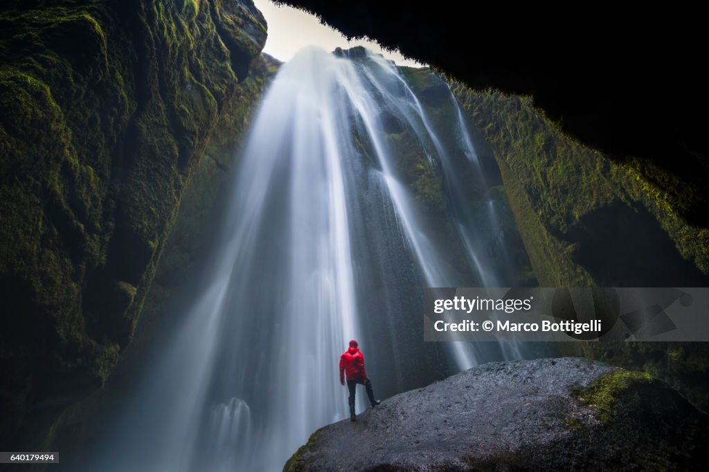 Man with red coast standing on top of a rock under Gljúfrafoss waterfall, Southern Iceland, Iceland, Northern Europe.