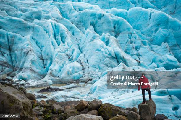 a man with red coat standing on top of a rock admiring the view of a glacier in skaftafell national park, eastern iceland, northern europe. - skaftafell national park stock pictures, royalty-free photos & images