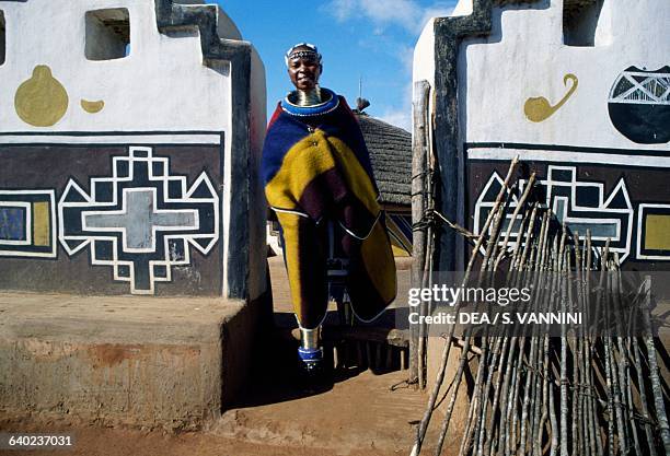Ndebele man wearing a traditional costume at the entrance to a house with decorated walls, South Africa.
