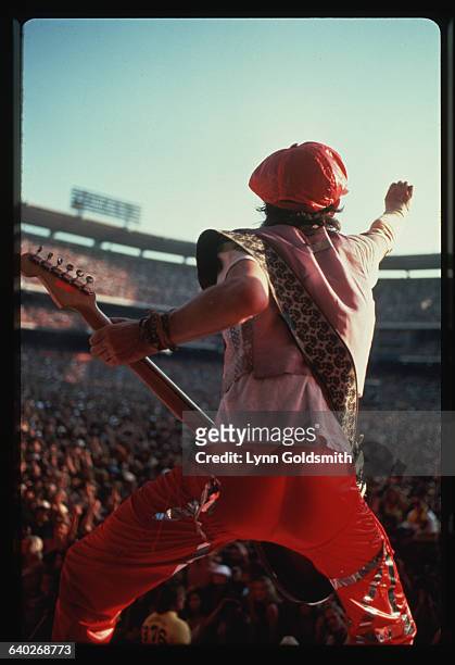 Mick Jagger, singer for the Rolling Stones, stands backstage holding an electric guitar and gesturing at a concert crowd.