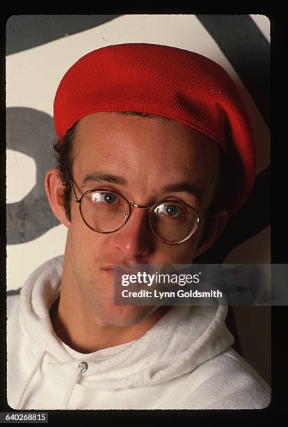 Close-up portrait of Keith Haring wearing red beret. He stands before a wall that he painted.