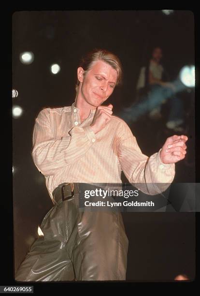 Sweaty David Bowie, in brown leather pants, strikes a dance pose on stage.