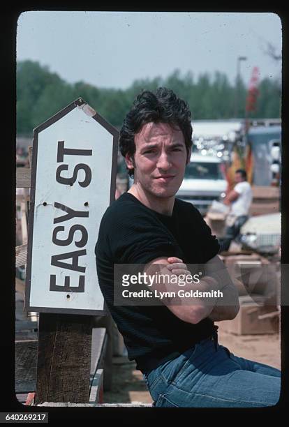 Rock and roll musician Bruce Springsteen poses outside against an arrow reading "Easy Street" and pointing up. Springsteen is wearing a black sweater...