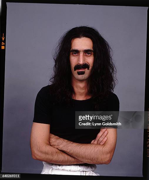 Studio portrait of musician Frank Zappa, standing alone in a black tee-shirt, with his arms folded. Undated color photo.