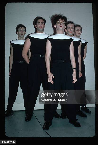 Picture shows the members of the band Devo posing together. They are standing in a pyramid style and wearing black outfits with white oval collars.