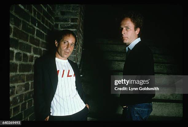 Portrait of Simon and Garfunkel. They are shown waist-up, posing against a brick wall. Photograph, 1981.