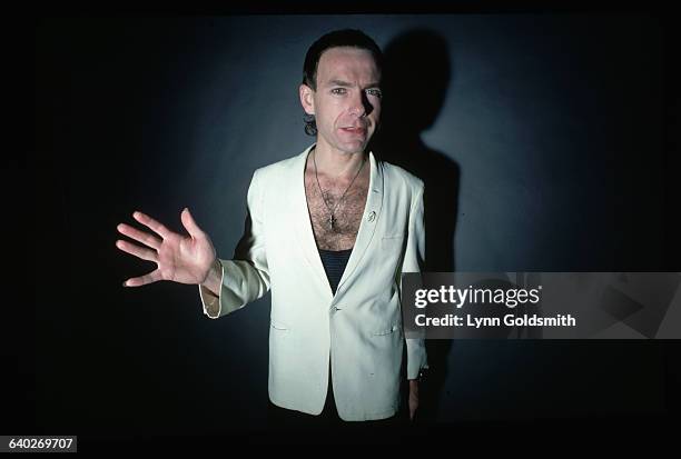 Picture shows guitar player, Robert Fripp, of the band "King Crimson", posing in a white jacket and low cut, black tank top. One hand is extended...