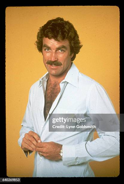 Actor Tom Selleck is shown standing in front of a yellow background in a studio portrait, buttoning his shirt.