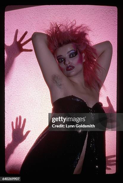 Picture shows German punk singer, Nina Hagan, posing in black sequins dress, pink hair and typical punk style makeup. Her hands are placed behind her...