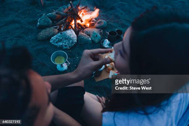 couple sharing food beside campfire on the beach at sunset - west asia stock-fotos und bilder