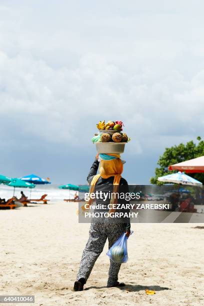 balinese woman carrying a bowl of fruits for sale on her head on kuta beach in bali - kuta stock-fotos und bilder