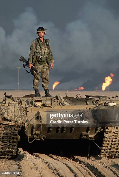 An American soldier stands on top of a destroyed Iraqi tank. Kuwaiti oil wells, ignited by Saddam's retreating forces, burn in the distance.