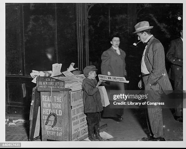 Year old newsboy sells a paper to a pipe-smoking businessman on Columbus Circle in New York City. His newsstand bears ads for his paper, the Herald,...