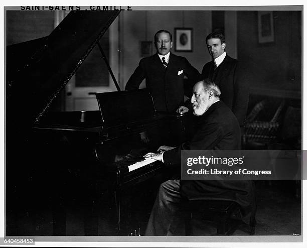 Composer Camille Saint-Saens plays the piano while two men look on.