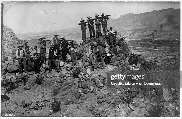 Mexican rebel group aims and shoots from a boulder overlooking a desert valley with factories. Mexico