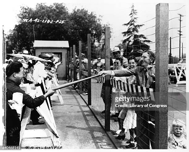 White American gives a baseball bat to an interned Japanese American, through a wire fence, at the Santa Anita Assembly Center at Santa Anita...