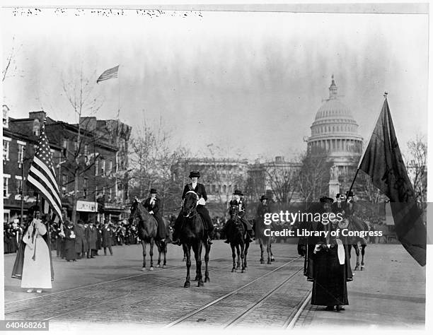 Some women march and carry flags, others ride horses in a suffrage parade on a street by the Capitol Building in 1913. Washington D.C., USA.