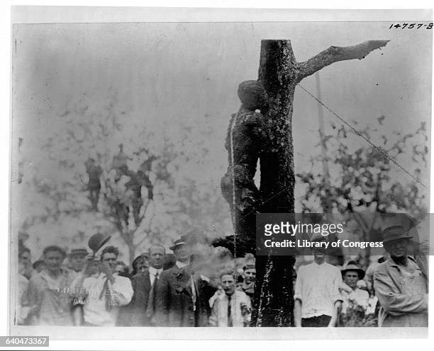 Crowd of people stands to watch the lynching by burning of Jesse Washington whose charred corpse leans chained to the trunk of a tree. Waco, Texas.