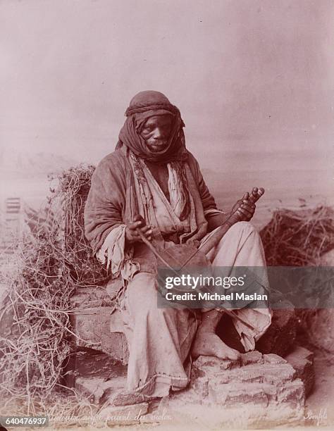 Bedouin man wearing a kaffiyeh sitting on a rock playing a stringed instrument with a bow. Jerusalem, ca. 1870s.