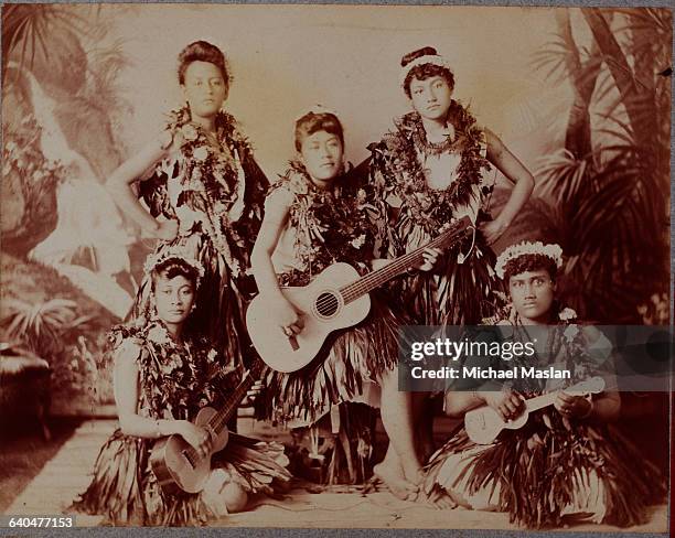 Five Hawaiian musicians and hula dancers, wearing traditional dress, hold guitars and ukuleles.