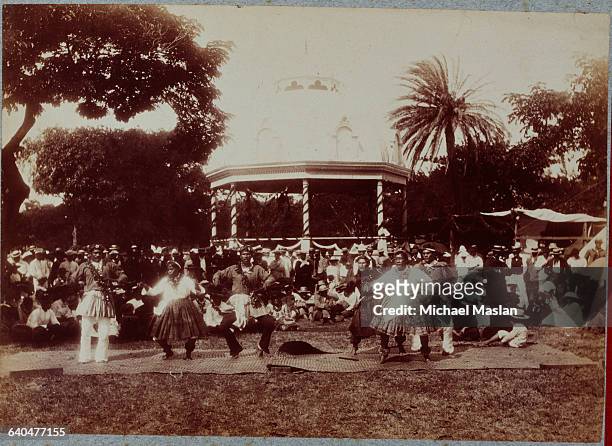 Male Hawaiian hula dancers, dancing at the King's palace in front of a large gazebo. They are wearing skirts, dancing on a mat before a crowd. Ca....