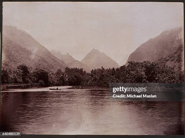 Man in a small, open boat on the Tautira River in Tahiti, ca. 1880. Steep hills thick with trees rise up behind the river.