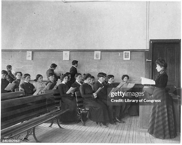 Teacher stands in front of young Native American students during class at the Carlisle Indian School, an institution dedicated to inducing Native...
