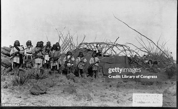 Armed men and children pose in front of a stick and blanket shelter at Geronimo's camp shortly before his surrender to General Crook, March 27, 1886.