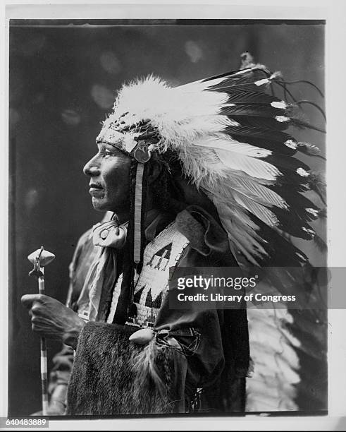 Ponca chief and Native American civil rights leader, Standing Bear, wearing a head dress and holding a ceremonial tomahawk, 1899-1900.