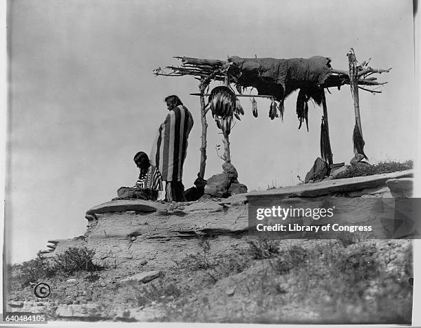 Two Piegan Blackfeet men next to burial scaffold with eagle feathers and animal hide, Montana, United States.