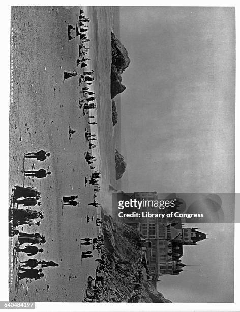 Crowd of people on the beach below the Cliff House enjoy the sun, the surf, and a view of Seal Rocks. The Gothic Revival style resort was built in...
