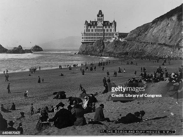 Crowd of people on the beach below the Cliff House enjoy the sun, the surf, and a view of Seal Rocks. The Gothic Revival style resort was built in...