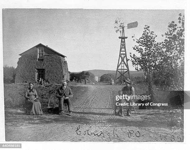 Four settlers gather in front of a sod house, with a windmill in the distance. | Location: Coburgh, Nebraska, USA.