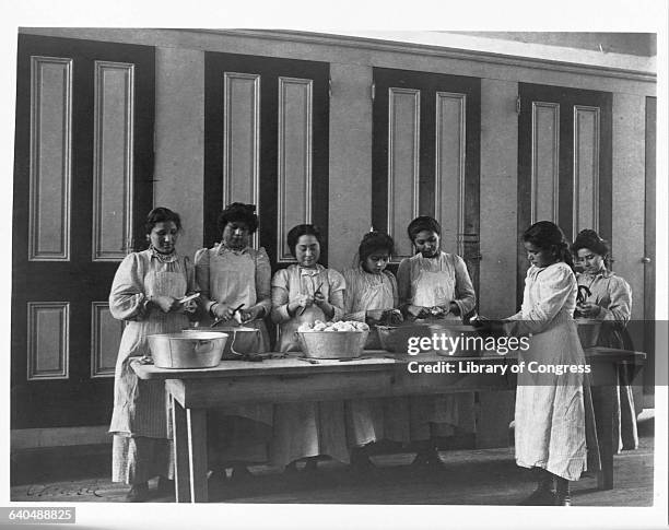 Young North American Indian women gathered around a table peel and slice buckets of apples in cooking class at the Carlisle Indian School, Carlisle,...