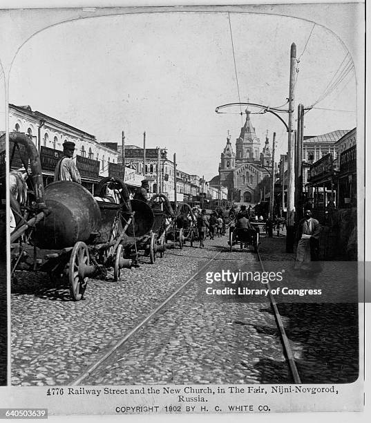 Carriages drive down Railway Streeet, with the New Church at the end, in The Fair, Nijini-Novgorod. Pedestrians walk beneath telegraph poles. |...