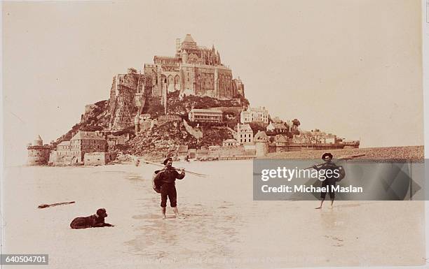 Two men stand on the beach on the south side of Le Mont-St. Michel, France. The abbey and city rises up behind them on the hill. Both men have poles...