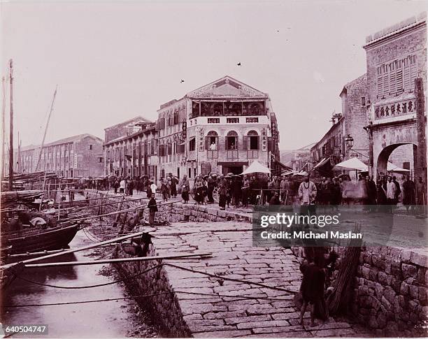 Crowd of Chinese people stand in front of a brick Chinese hotel on the waterfront of Macao, a Portugese colony on the coast of China. A row of junks...