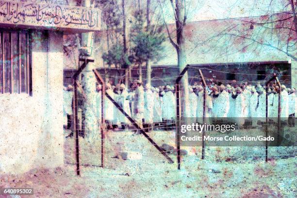 View through barbed wire fencing of a group of men dressed in white robes and headbands, standing with fists raised in a large courtyard in Iran,...