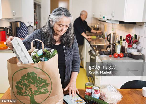 senior woman removing grocery from shopping bag while using digital tablet in kitchen - quitar fondo de imagen online fotografías e imágenes de stock